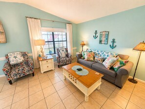 Ein Wohnzimmer mit einem gemusterten Sessel, einer Couch mit floralen Kissen, einem Holztisch und maritimer Wandgestaltung.