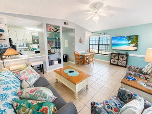Ein Wohnzimmer mit Essbereich, Deckenventilator, Fernseher mit Blick auf den Strand und einer Küche im Hintergrund.