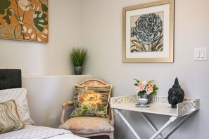 Una habitación con una cama, una silla con un cojín floral, una mesita con una estatua de Buda, un grabado botánico enmarco y una planta en maceta.