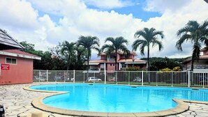 Una piscina con solarium de piedra, rodeada por una valla blanca, palmeras y un edificio de techo rojo.