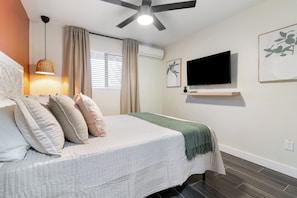 Ein Schlafzimmer mit einem Bett, einem Deckenventilator, einem Fernseher und einem Fenster mit Vorhängen.