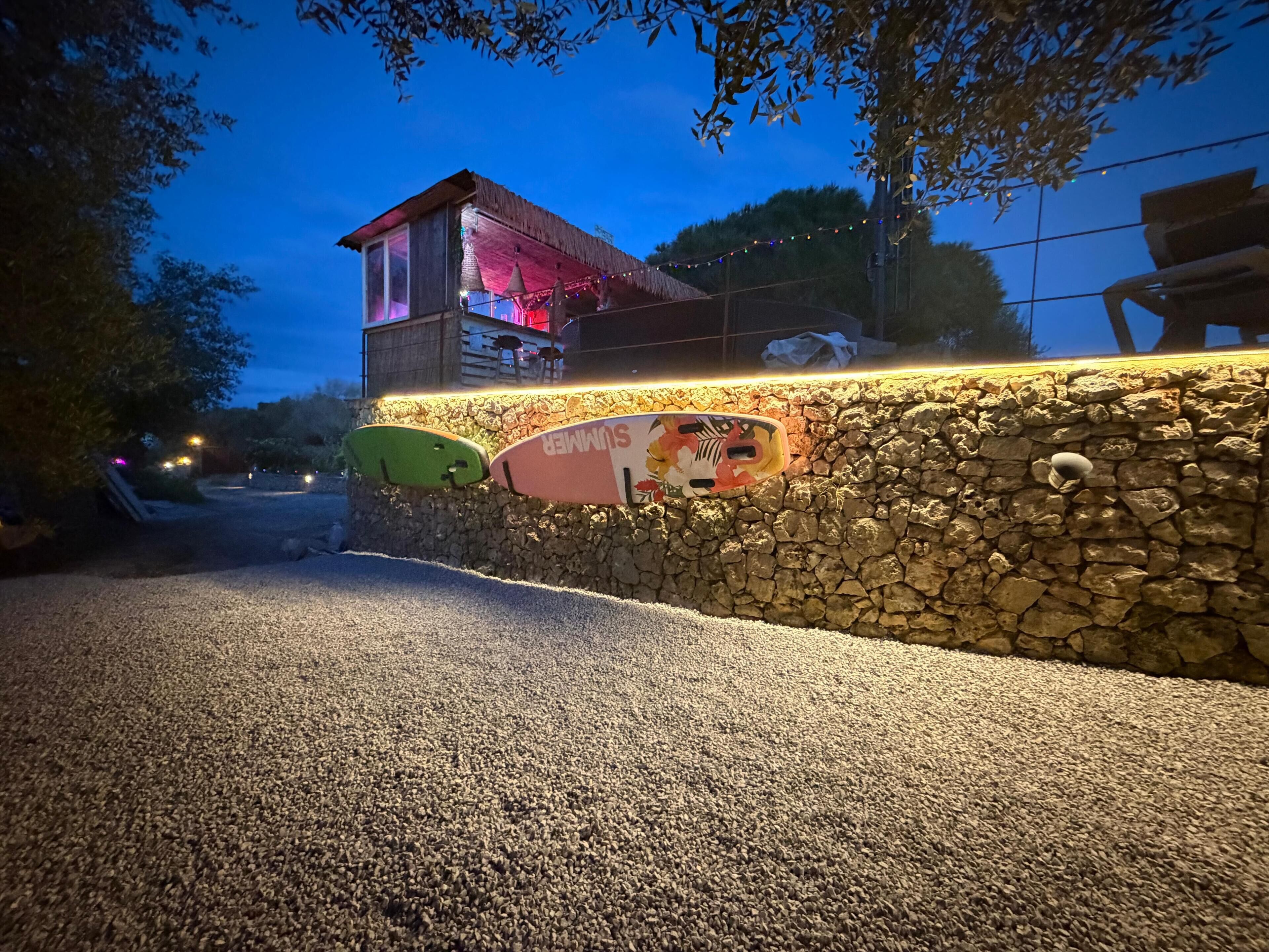 Un muro de piedra con tablas de surf, un edificio con techo de paja y un camino de grava.