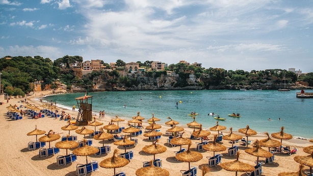 Une plage avec de nombreux parasols et des chaises longues, une tour de surveillance pour les sauveteurs, et des bâtiments en arrière-plan.