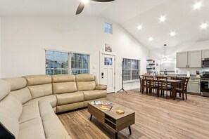 Un salon moderne comprenant un canapé d’angle, une table à manger avec des chaises et un coin cuisine avec des placards et des appareils électroménagers.
