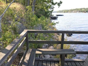 A wooden deck with a railing overlooking a calm lake, surrounded by trees and rocks. - Lovely log house with a view of the sea (Figeholm)
