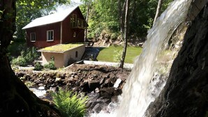 Uma cabana rústica com telhado inclinado, uma pequena construção anexa e uma cascata ao fundo.