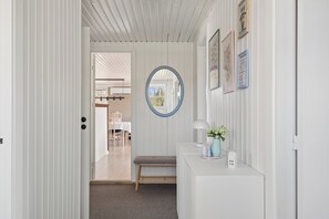 Un couloir avec une porte blanche, un banc, un miroir rond et une petite table sur laquelle se trouvent une lampe et un vase.
