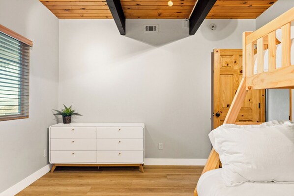 Un dormitorio con literas, un aparador blanco, techo de madera y una ventana con persianas.