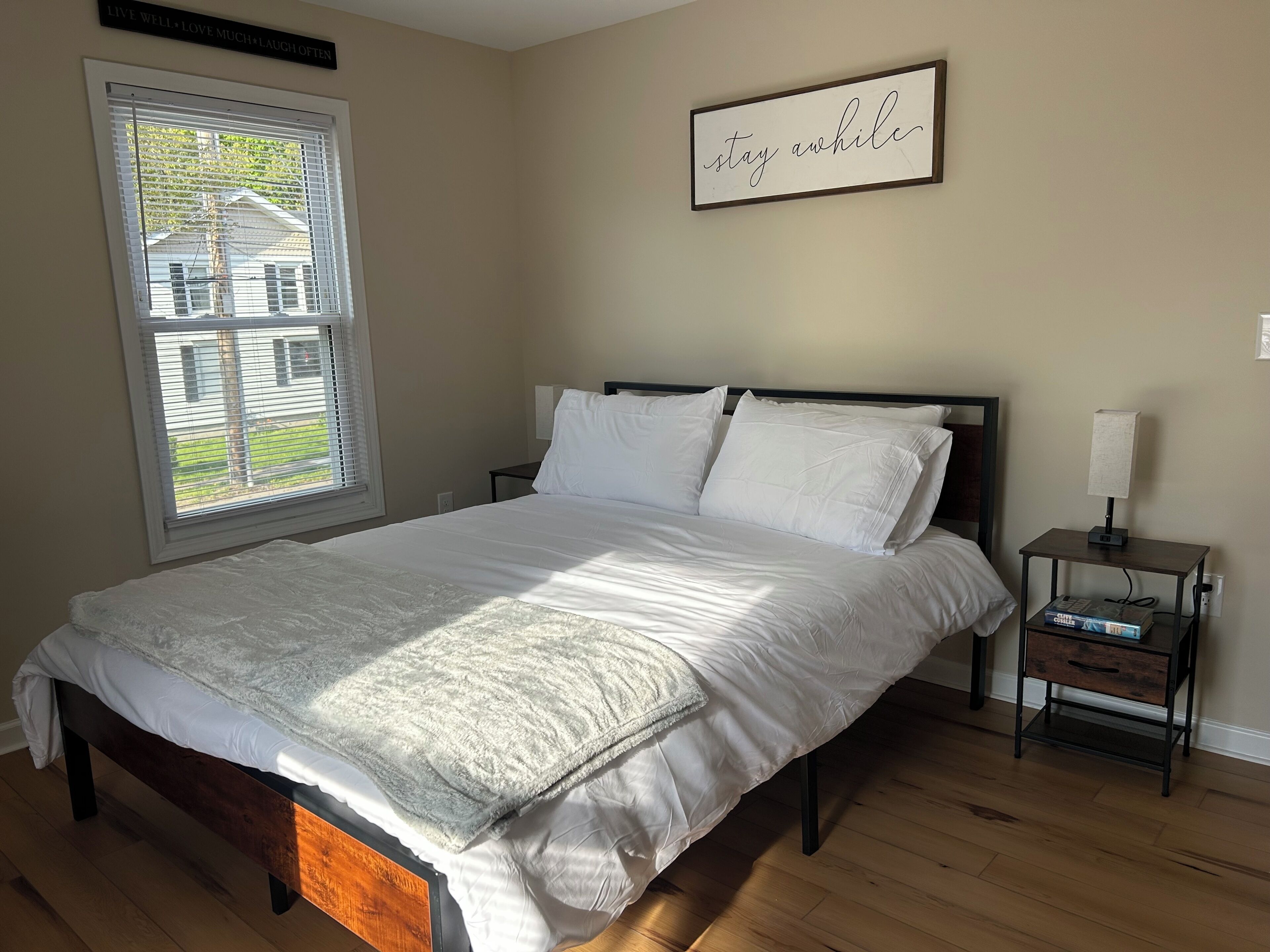 A neatly made bed with white linens, a bedside table with a lamp, and a window with blinds.
