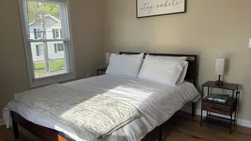 A neatly made bed with white linens, a bedside table with a lamp, and a window with blinds.