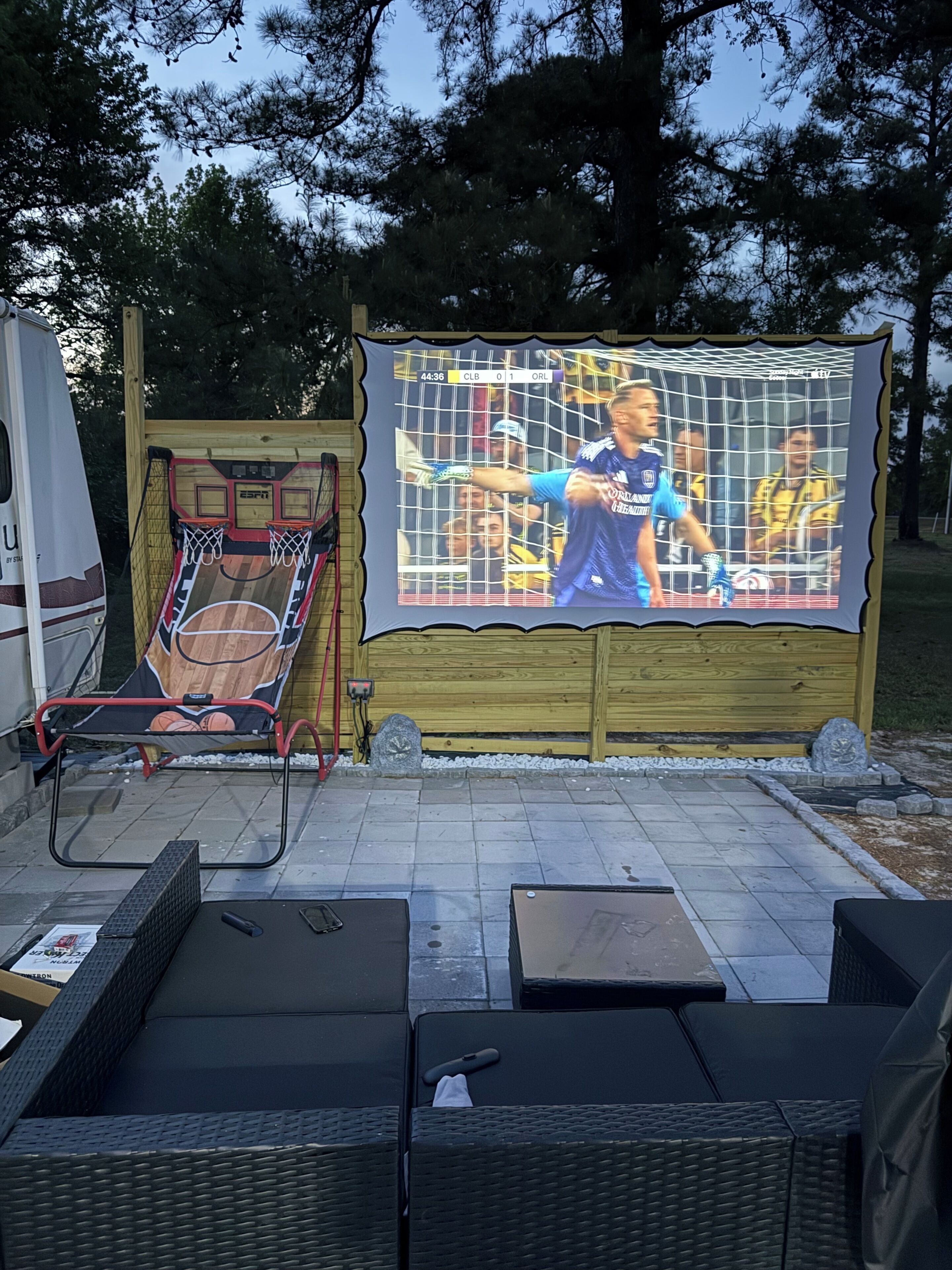 Un espace extérieur aménagé avec des sièges, un panier de basketball et un grand écran diffusant un événement sportif.