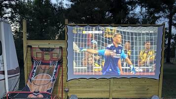 Un espace extérieur aménagé avec des sièges, un panier de basketball et un grand écran diffusant un événement sportif.