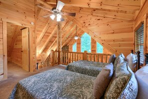 Camera da letto in stile baita con soffitto spiovente, ventilatore a soffitto, un letto con biancheria a motivi decorativi e una finestra vista alberi.