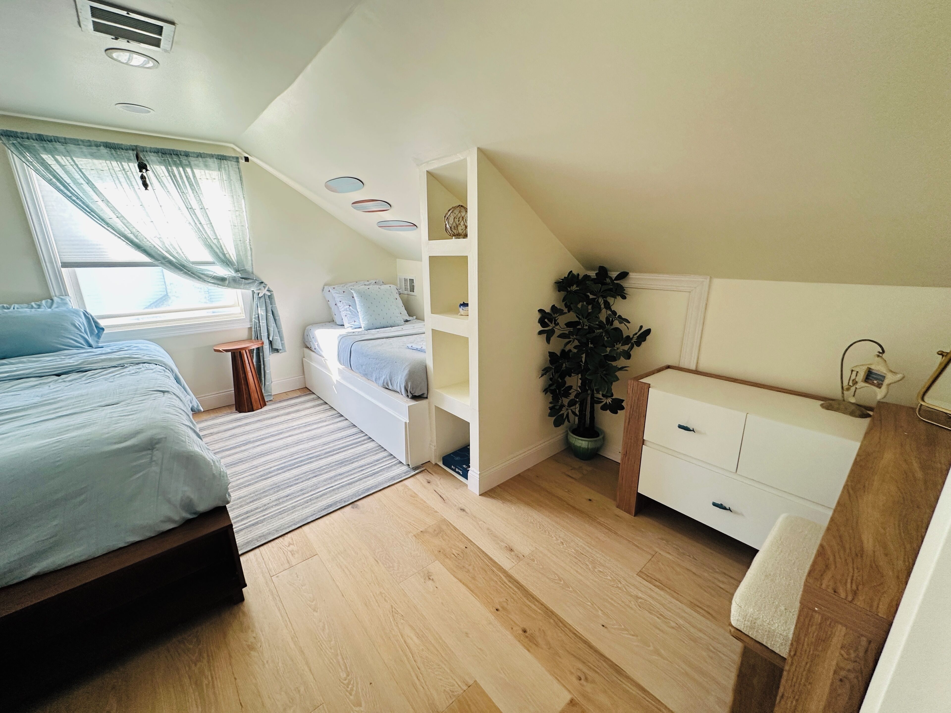 A bedroom with a bed, bedside table, dresser, potted plant, and a window with curtains.