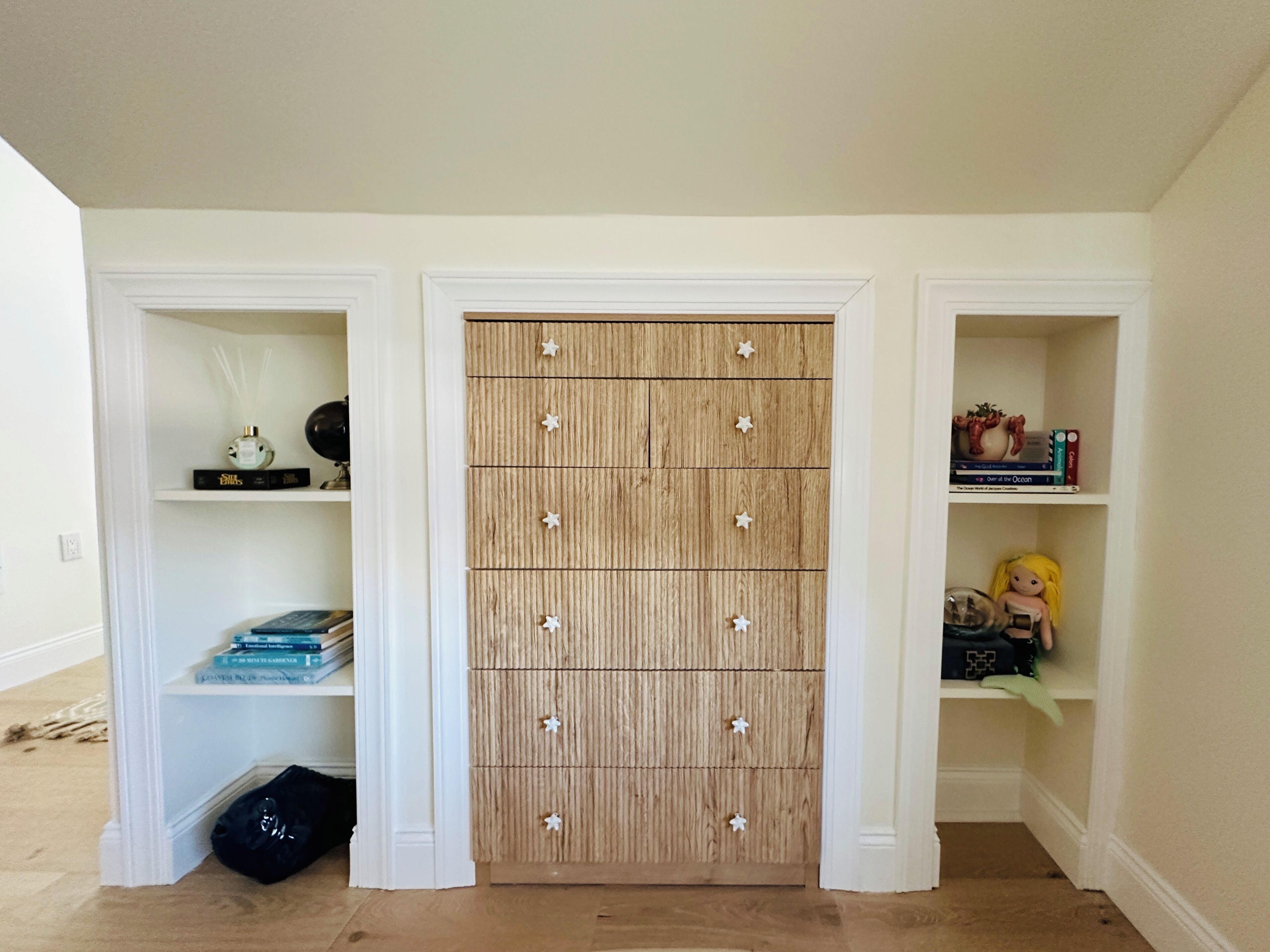 Een kamer met een houten kast, twee witte planken met boeken en decoratieve spullen, en een zwarte tas op de vloer.