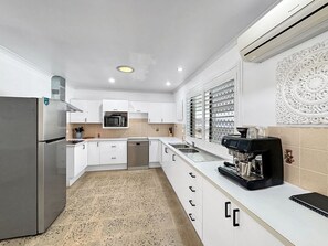 Una cocina moderna con armarios blancos, un refrigerador de acero inoxidable, lavavajillas, cafetera y ventana con persianas.