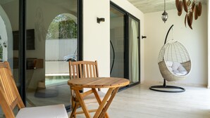 Uma cadeira e mesa de madeira numa varanda, com uma cadeira suspensa, portas de vidro e vista para uma scooter.