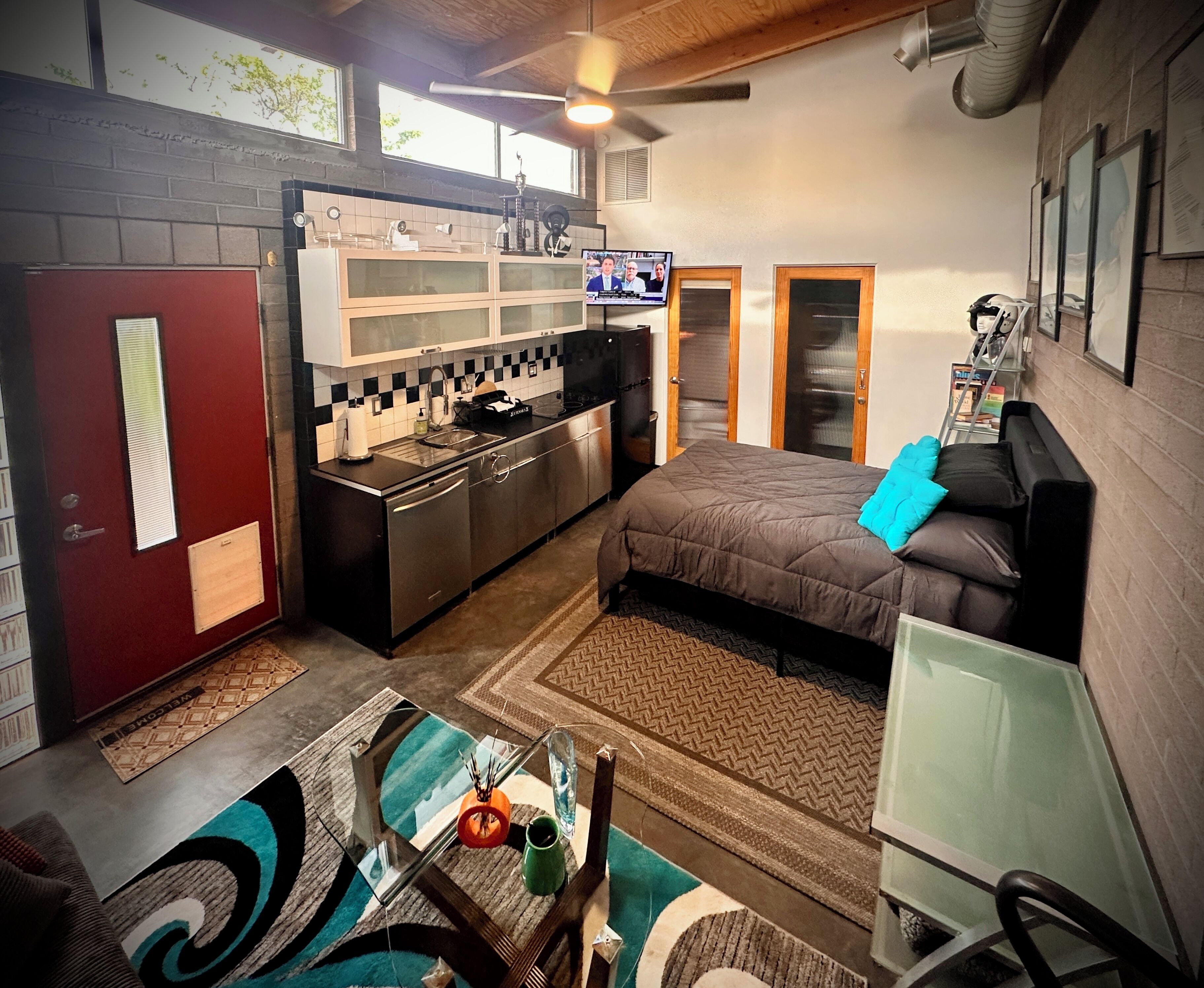 A compact room with a kitchenette, a bed with a blue pillow, a dining table with chairs, and a TV mounted on the wall.