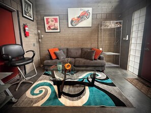 Ein Zimmer mit einem grauen Sofa, einem schwarzen Sessel, einem Glastisch, einem gemusterten Teppich, einem Motorradbild und einem Feuerlöscher.