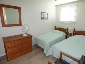 A bedroom with two single beds, a wooden dresser, a mirror, a fan, a bedside table with a lamp, and a window with blinds. - Bright & Welcoming 2BR Near Beach, Boardwalk & Local Attractions (Ocean City)