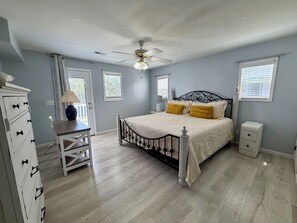 Ein Schlafzimmer mit Bett, Schminktisch, Nachttisch, Lampe, Deckenventilator und Fenstern.