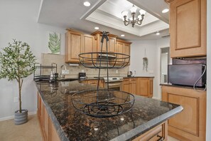 A kitchen with wooden cabinets, a granite countertop, a three-tiered wire basket, a potted plant, and a modern chandelier. - Palm Harbor Family Retreat, 2 Kayaks, Air Hockey, Foos Ball, Fishing, Boat Lift (Palm Harbor)