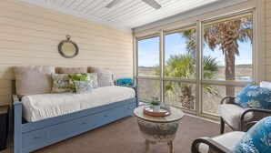 Un dormitorio con cama, ventilador de techo, ventana con vista a palmeras y una mesita con una planta.