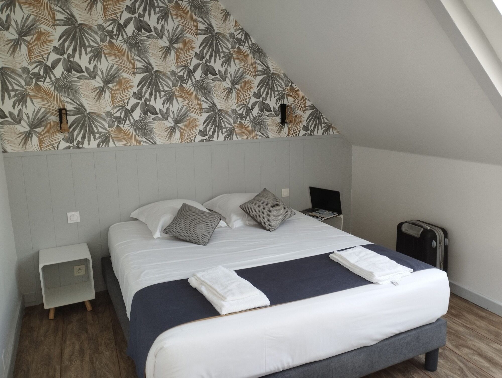 A neatly made bed with pillows, a bedside table, a laptop, and a suitcase in a room with a sloped ceiling and a leaf-patterned wall.