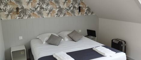 A neatly made bed with pillows, a bedside table, a laptop, and a suitcase in a room with a sloped ceiling and a leaf-patterned wall.