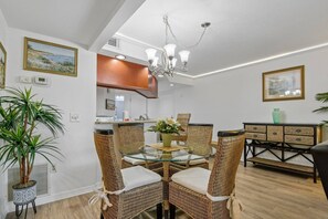 Ein Essbereich mit Korbstühlen, einem Tisch mit Glasplatte, einer Pendelleuchte und einer Küche im Hintergrund.