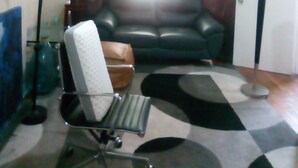 Un salon avec un canapé en cuir noir, un fauteuil de bureau en verre, un tapis à motifs, un lampadaire et un tableau encadré accroché au mur.