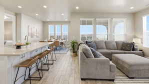 A modern living room with a grey sofa, a dining area with a long table, and a kitchen with a bar.