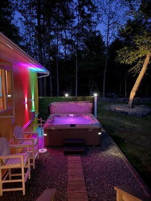 Un jacuzzi con terraza de madera y sillas, rodeado por un seto y árboles.