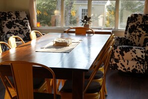 A dining area with a wooden table, chairs, and a window with a view of buildings and trees. - Nemandua Catskills: Hot Tub+Fire Pit+Pool Table+EV (Lanesville)