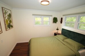A bedroom with a bed, two windows, a nightstand, and a wall-mounted plant. - Nemandua Catskills: Hot Tub+Fire Pit+Pool Table+EV (Lanesville)