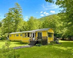 A yellow cabin with a porch, surrounded by lush greenery and trees. - Nemandua Catskills: Hot Tub+Fire Pit+Pool Table+EV (Lanesville)