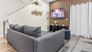 A modern living room with a grey sofa, a flat-screen TV, a wooden panel wall, and a white curtain. - 4BR Disney Area Home and Resort, Themed Rooms, Arcade Games, Gym and Pool. (Davenport)