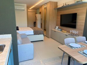 Ein kompaktes Hotelzimmer mit Bett, Küchenzeile, Essbereich und einem an der Wand montierten Fernseher.