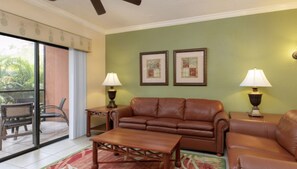 A living room with a brown leather sofa, a wooden coffee table, two side tables with lamps, and a large sliding glass door leading to a balcony. - Spacious 1BR Deluxe Villa w/ Full Kitchen, Balcony & Jetted Tub - 800 Sq Ft (Orlando)