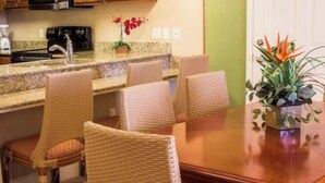 A dining area with wicker chairs, a wooden table, and a kitchen in the background. - Spacious 1BR Deluxe Villa w/ Full Kitchen, Balcony & Jetted Tub - 800 Sq Ft (Orlando)