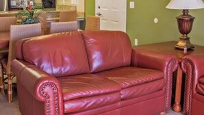 A red leather sofa in a room with a green wall, a wooden table with a lamp, and a kitchen area in the background. - Spacious 1BR Deluxe Villa w/ Full Kitchen, Balcony & Jetted Tub - 800 Sq Ft (Orlando)