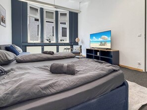 Ein Schlafzimmer mit Bett, Fernseher, Fenster mit Rollläden und Blick auf den Strand.