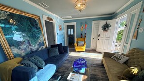 A living room with a blue sofa, a yellow armchair, a glass coffee table, and a large abstract painting. - 0.5 Mile Walk To Sofi - Art Deco Beach Cottage - Entire Home (Inglewood)