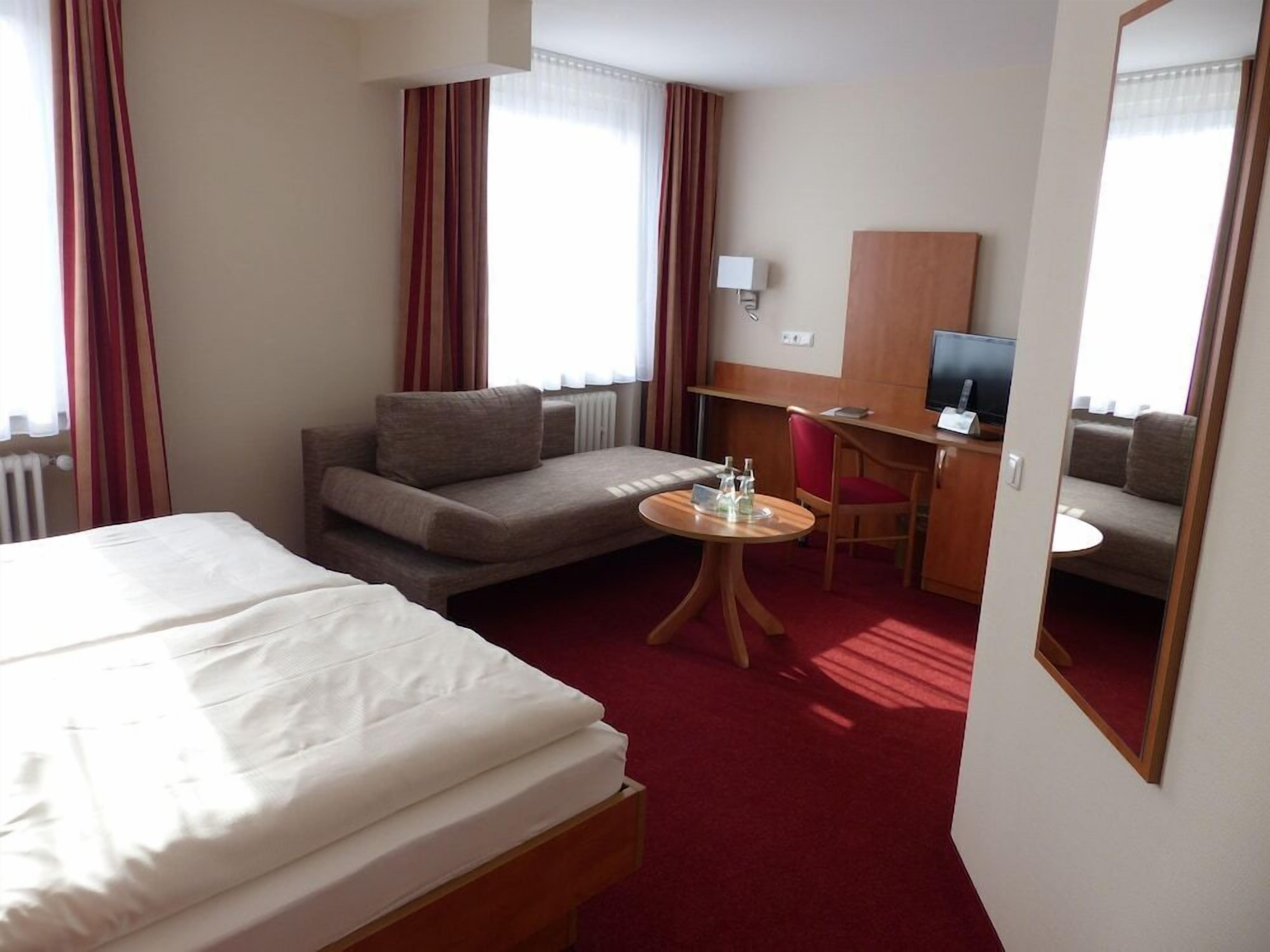 Ein Hotelzimmer mit Bett, Sofa, Schreibtisch mit Stuhl, Fernseher und Spiegel.