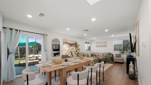 A dining area with a wooden table set for six, surrounded by white chairs. There is a large window with curtains, a sofa, and a TV stand with decorative items. - King Bed, Garage, Gated Backyard, 4BR/2Bth (El Cajon)