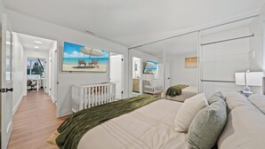 A bedroom with a large bed, a crib, a white wall with a beach painting, and a view of the living area. - King Bed, Garage, Gated Backyard, 4BR/2Bth (El Cajon)
