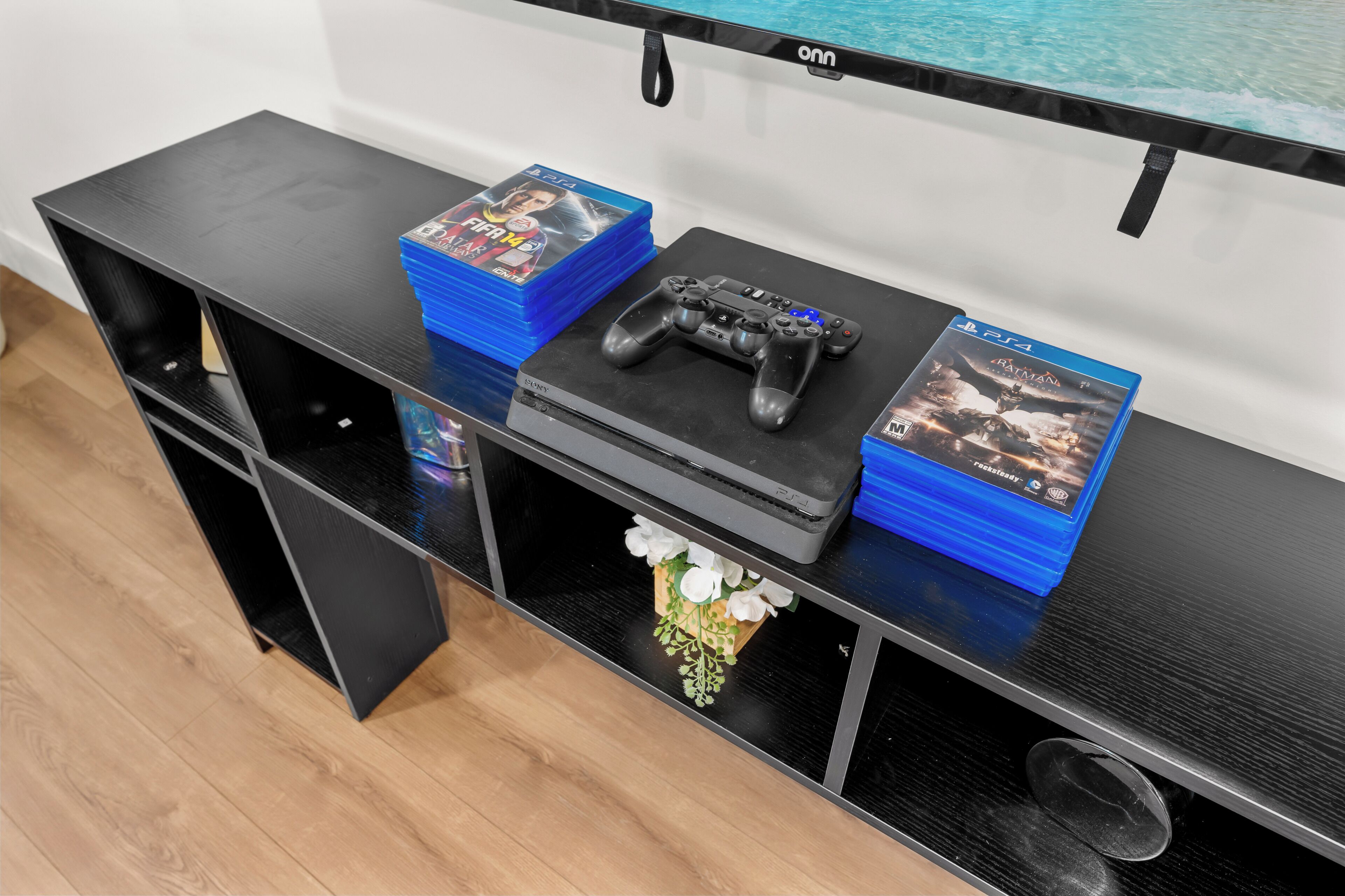 Eine schwarze Unterhaltungsanlage mit einer PlayStation 4 Konsole, einem Controller und mehreren Spielehüllen, darunter FIFA 21 und Batman Vengeance.