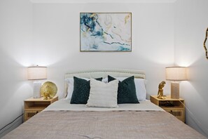 A neatly made bed with a headboard, two bedside tables with lamps, and a wall-mounted abstract painting. - King Bed, Garage, Gated Backyard, 4BR/2Bth (El Cajon)