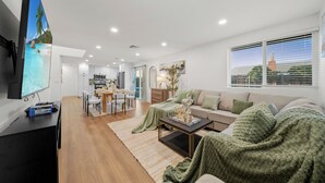 A modern living room with a flat-screen TV, a sofa, a coffee table, and a dining area with a table and chairs. - King Bed, Garage, Gated Backyard, 4BR/2Bth (El Cajon)