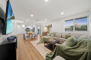 A modern living room with a flat-screen TV, a sofa, a coffee table, and a dining area with chairs and a table. - King Bed, Garage, Gated Backyard, 4BR/2Bth (El Cajon)